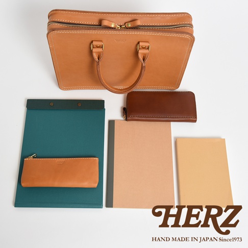 職人手作皮革手提商務公事包，淺棕色皮革，配有頂部提把、拉鍊開合及金色五金配件。旁邊擺放著一個深棕色長夾、一個淺棕色筆袋、一本綠色資料夾、一本卡其色筆記本和一本淺黃色筆記本。