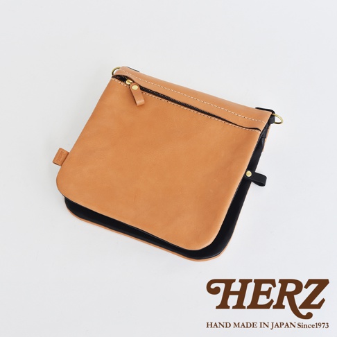 職人手作皮革翻轉雙色直式斜背包，以鞣製皮革製成，呈現自然的棕褐色調，搭配黑色包邊與內裡。包款設有拉鍊開口、皮質拉環，以及用於連接背帶的金屬環。