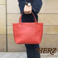 紅色職人手作皮革極簡托特包，配有兩個短提把和兩個金色铆釘，包身呈現細膩的皮革紋理，由穿著牛仔褲和深藍色毛衣的人士手持。