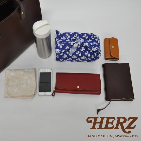 深棕色皮革直式托特包，旁邊擺放著不鏽鋼保溫瓶、印有貓咪圖案的藍色風呂敷、米白色繡花手帕、白色手機、紅色皮革長夾及深棕色皮革筆記本。