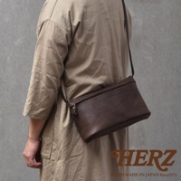 深棕色皮革製成的斜背包，由人斜背於身，包款有拉鍊開口，右下角有HERZ字樣及HAND MADE IN JAPAN Since1973字樣