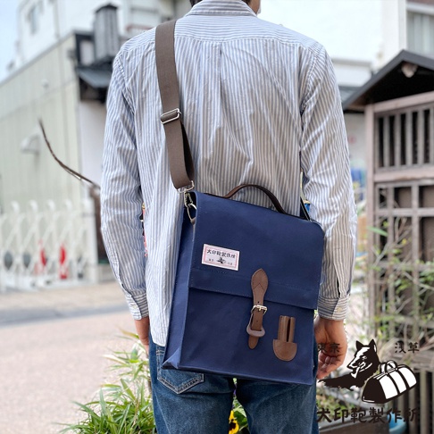 身著條紋襯衫與牛仔褲的男子，斜挎一個深藍色純棉帆布直式側背包，包包上有皮革束帶與釦環，並附有提把，呈現簡約風格。
