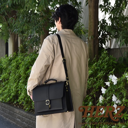 職人手作黑色皮革三用單扣商務後背包，展示於一位身穿米色外套的男士肩上，配有可調節的黑色皮革肩帶和金色扣環。