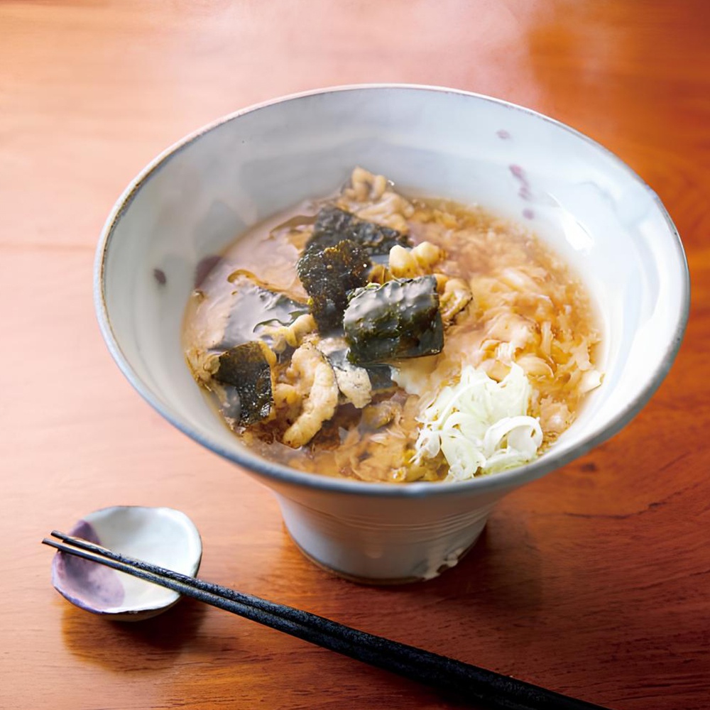 淺色陶瓷碗中盛有海苔天婦羅餅乾，湯色呈琥珀色，餅乾塊狀，點綴著深綠色海苔和白色蔥花。一雙黑色的筷子放在旁邊的紫色小碟上。