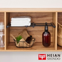 展示一個黑色的伸縮縫隙置物架，架上擺放著疊好的白色毛巾、一個裝有麻布和綠植的黑色線籃，以及一個琥珀色噴霧瓶。置物架安裝在木紋的層架空間內。