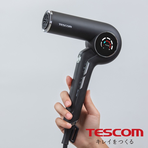 一手拿著一支黑色TESCOM吹風機，吹風機上顯示有溫度指示燈，下方印有TESCOM的Logo及日文文字。