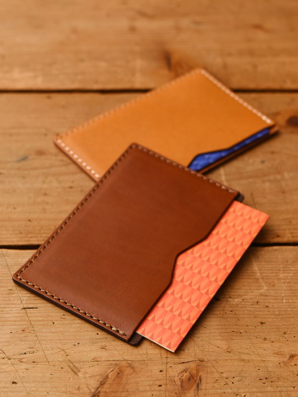 兩款職人手作皮革文件夾，一款為棕褐色皮革，另一款為淺棕色皮革，均採用手工縫線。淺棕色皮革夾內露出部分藍色內襯，棕褐色皮革夾則插有一張橘色幾何圖案卡片。