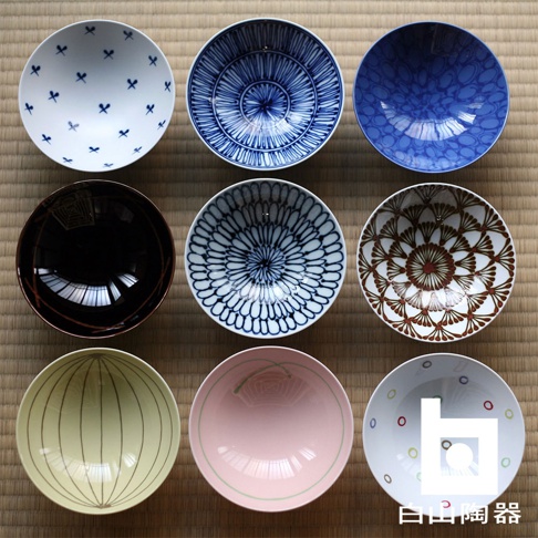 九個陶瓷平茶碗，展示了多種設計：白色碗身飾有藍色蝴蝶結圖案；藍白相間的放射狀紋理；藍色碗身帶有圓形圖案；黑色碗身；藍白相間的花卉圖案；紅棕色與白色相間的扇形紋理；黃色碗身飾有細長條紋；粉色碗身飾有綠色線條；白色碗身飾有彩色圓點。