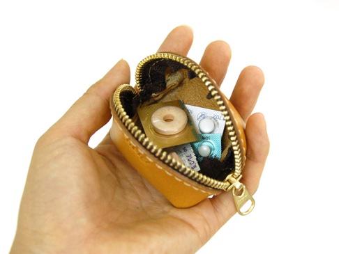 手持一只 tan 色皮革貝殼造型零錢包，拉鍊敞開，內部可見藥丸藥片，零錢包由皮革製成，內襯為深棕色。