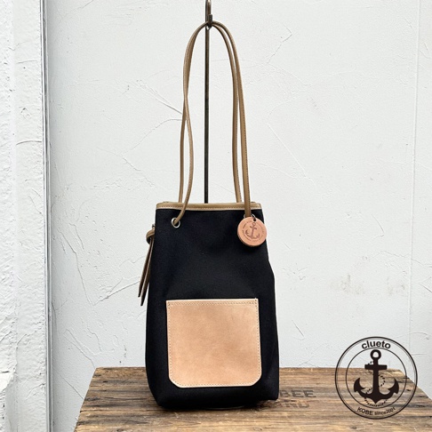 一個黑色帆布束口袋肩背包，配有咖啡色皮革提把和方形皮革前袋，包上掛有船錨造型的吊牌，背包底部放在木製檯面上。