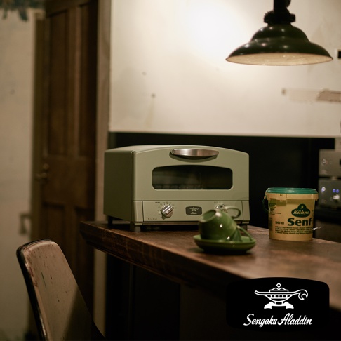 淺綠色復古多用途烤箱，配有銀色提把和兩個烤網，左側有兩個旋鈕，右側有品牌標誌，前方有玻璃視窗，烤箱置於木質檯面上，旁邊有一個小綠色杯子和碟子，以及一個黃色罐子。