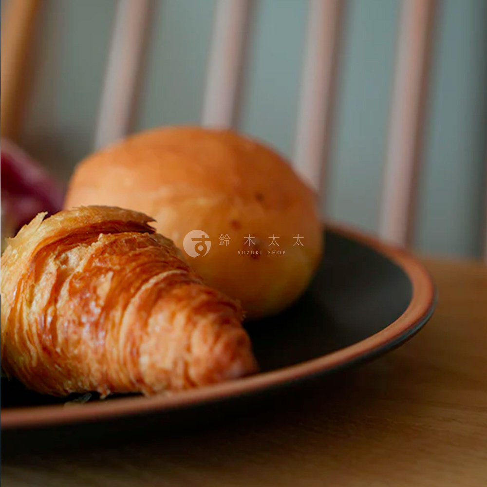 PURE 素燒百搭陶瓷主菜盤 M，黑色盤身搭配焦糖色盤緣，盤中放著一個可頌麵包和一個圓形麵包。