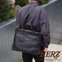 男士身背黑色皮革托特包，包包有雙提把和可拆卸肩帶，採用厚實皮革製作，設計簡約。