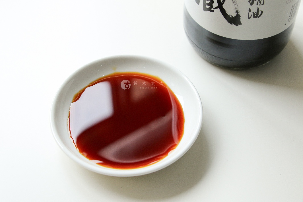 白瓷碟中盛有深褐色醬油，碟旁置有標有黑色漢字「秘藏醬油」的黑色瓶身，瓶頸處標有「SUZUKI SHOP」字樣。