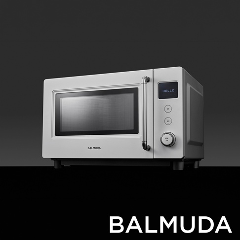 這是一款 BALMUDA 品牌的白色微波爐，採橫向長方體設計，側面設有顯示螢幕與旋鈕控制面板，外觀簡約現代，正中央飾有金屬感手把，整體呈現於純灰背景上，下方標註 BALMUDA 字樣。
