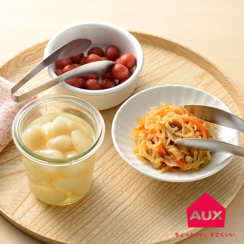 一對銀色金屬夾子，夾著日式漬物，旁邊有一個裝有醃漬小洋蔥的玻璃罐，以及一個裝有梅子的白色圓形碗，皆放置於木質托盤上。