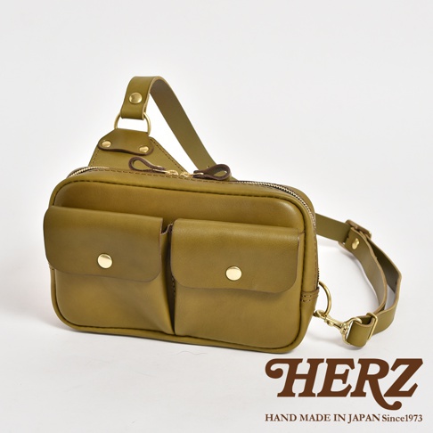 橄欖綠皮革雙口袋斜背包，設有可調節肩帶、頂部拉鍊開口，以及兩個帶有金色按扣的前袋。