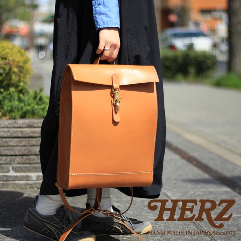 棕褐色皮革製成的紙袋風後背包，配有金色金屬扣環和皮革提把，展現簡約設計。