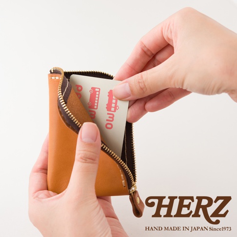 一雙手正拿著一個日本HERZ職人手作的棕色皮革迷你零錢包，拉鍊呈現半開狀態，其中一隻手正在將一張印有粉紅色圖案的卡片放入錢包內的夾層中，畫面右下角帶有HERZ品牌字樣。