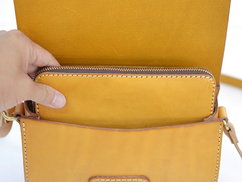 職人手作皮革寬扣帶馬鞍包，明亮的橘黃色皮革，包內可見一個拉鍊長夾，由細緻縫線固定。