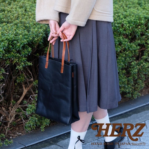 一位穿著淺色外套與深色百褶裙的女性，手中提著一個由日本職人品牌HERZ手工製作的黑色皮革直式托特包，包身設計簡約，配有棕色皮革提把，整體風格溫潤自然。