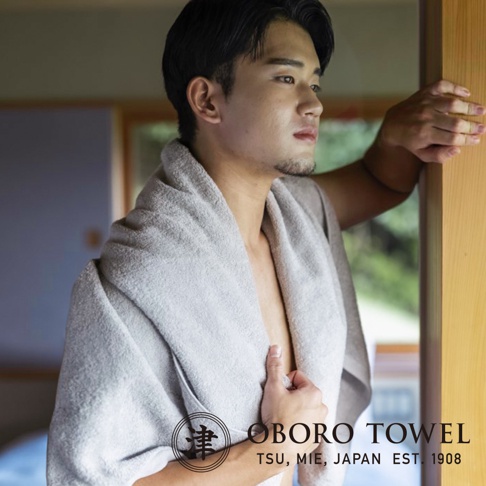 一位年輕男性身披淺灰色細纖浴巾，浴巾質感柔軟，呈現摩登復古風格，搭配品牌 Logo。