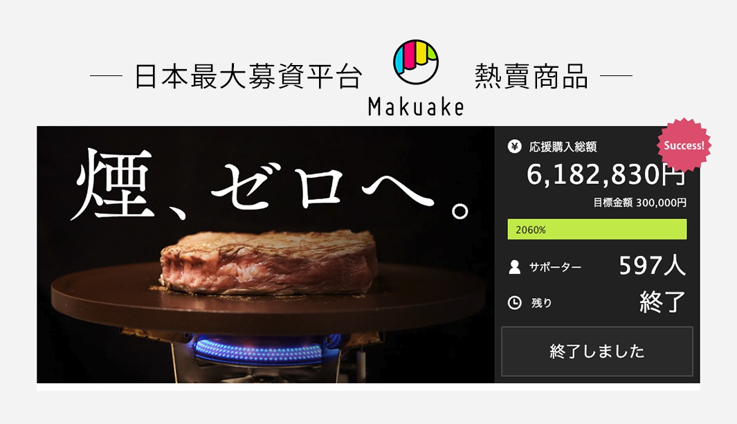 一塊厚切牛排放在一個圓形、深棕色的烤盤上，烤盤下方是藍色的火焰。烤盤上方有日文文字「煙、ゼロへ。」。右側顯示募資平台 Makuake 的成功訊息，包含募資金額、目標金額、支持人數及狀態。