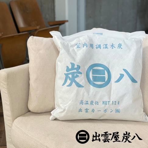 這張圖片顯示一個方形的出雲屋炭八室內調濕木炭，包裝為白色不織布，袋面印有藍色的炭八品牌標誌與產品資訊，並擺放在淺色布質沙發椅上。