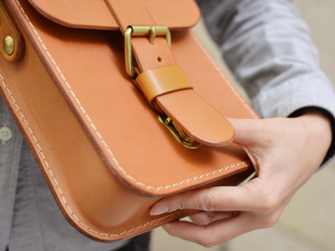 職人手作皮革經典皮扣單肩包，採用天然皮革材質，呈現溫潤的焦糖棕色。包身線條簡潔，邊緣有手工縫線裝飾，搭配黃銅色金屬皮扣，整體風格經典耐看。