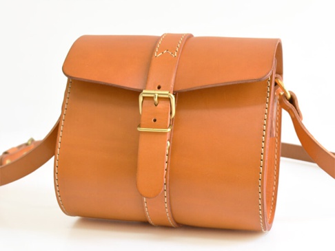 一只棕褐色牛皮製成的單肩包，帶有黃銅皮扣和肩帶，使用縫線裝飾。