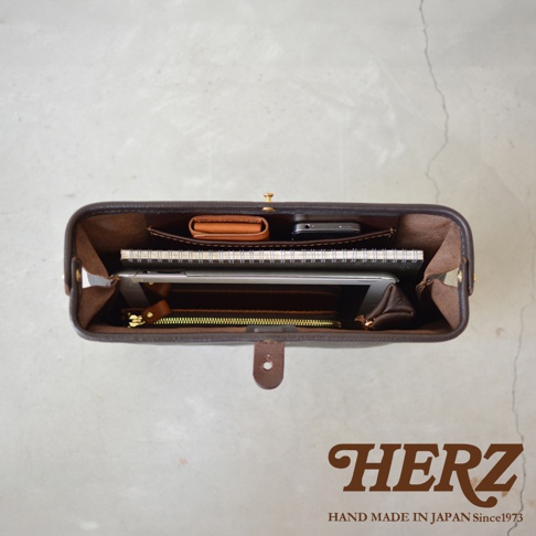 職人手作皮製Organ手拿包，深棕色皮革，內部裝有筆記本、平板電腦、錢包和手機，展現包款的收納功能。