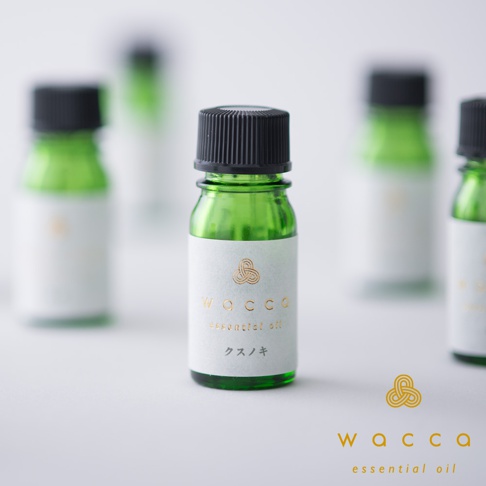 綠色玻璃瓶裝的吉野檜木精油，瓶蓋為黑色，瓶身貼有白色標籤，標籤上有金色品牌標誌、WACCA、essential oil 及日文「クスノキ」。背景有數瓶同款精油模糊呈現。