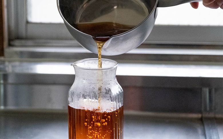 一個不鏽鋼雪平鍋正在將琥珀色液體倒入一個帶有條紋的透明玻璃罐中。