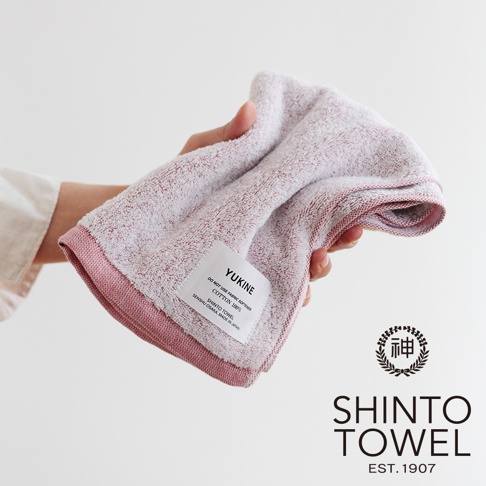 手持一條淡粉色、蓬鬆柔軟的 YUKINE 雪中漫步有機棉毛巾，毛巾表面帶有細緻的紋理，邊緣處有較深的粉色滾邊，掛有印有品牌名稱及產品資訊的白色標籤。