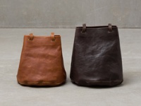 兩個職人手作皮革抽繩包，一個為淺棕色，一個為深棕色，皆為圓底桶狀設計，配有短提把。