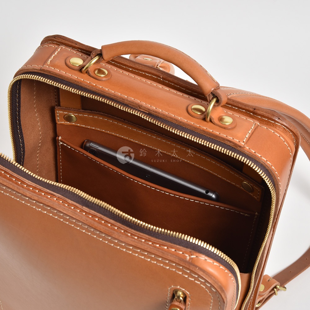 一個棕色皮革製成的職人手作商務電腦後背包，頂部有手提把，主隔層打開，內部有手機或平板電腦收納袋。