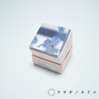 透明壓克力立方體中封存著一朵藍色翠雀花，下方為粉色紙盒包裝，盒上標示「デルフィニウム Siberian larkspur」。