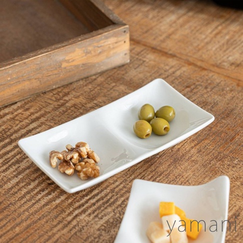 白色陶瓷小菜盤，有兩個分隔區，左側裝有核桃，右側裝有綠橄欖。盤子呈長方形，邊緣略微彎曲。背景是木紋桌面和一個木盒。