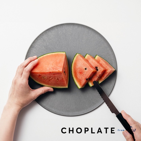 一個灰色圓形CHOPLATE兩用砧板盤，盤上有切好的西瓜塊，一隻手拿著其中一塊西瓜，另一隻手持刀準備切下一塊。