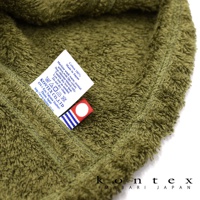 綠色毛巾特寫，展示其柔軟材質，左側有白色洗標，印有「KONTE X CO., LTD」字樣及產品說明，下方附有藍白條紋與紅色圓點的品牌織帶。