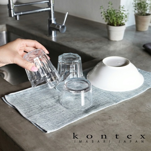 一張淺藍色棉麻混紡的 KONTEX 拭巾平鋪在水泥檯面上，上方擺放著兩個玻璃杯及一個白色碗，一隻手正輕輕扶著其中一個玻璃杯，背景處有水龍頭與盆栽，呈現簡約清新的廚房生活場景。