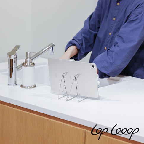 銀色金屬伸縮式平板支架，置於白色檯面上，支架上的平板電腦背面朝向觀看者，一旁有銀色金屬水龍頭和白色皂液器。