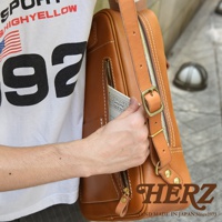 職人手作皮革長方型後背包，採用鞣製皮革材質，呈現自然的棕褐色光澤。背包設有多個拉鍊口袋，其中一個前置拉鍊口袋可見一本小筆記本。肩帶配有金屬釦環，可調節長度。