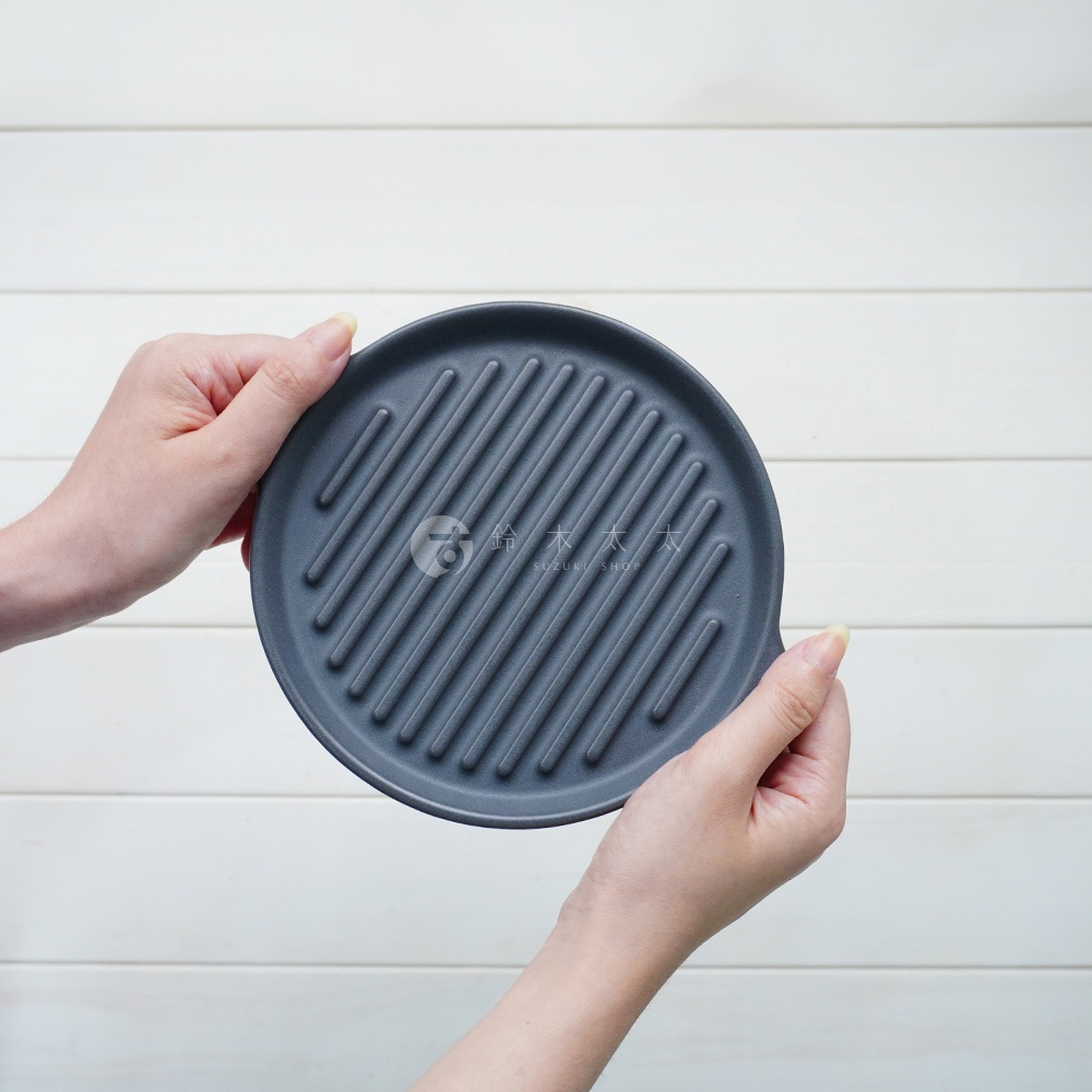 灰色陶製圓形烤盤，表面有規則的波浪紋路，由雙手持握，背景為白色木紋。
