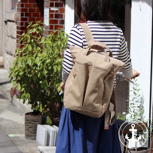 身穿藍白條紋上衣的女性，背著一個米色高島帆布後背包，背包有多個外袋和提把，整體為簡約設計。