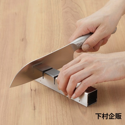 一雙手正使用 ESSYO 越匠系列可拆式不鏽鋼雙滾輪磨刀器，將一把銀色金屬刀刃的廚刀在木質桌面上的磨刀器上進行研磨。