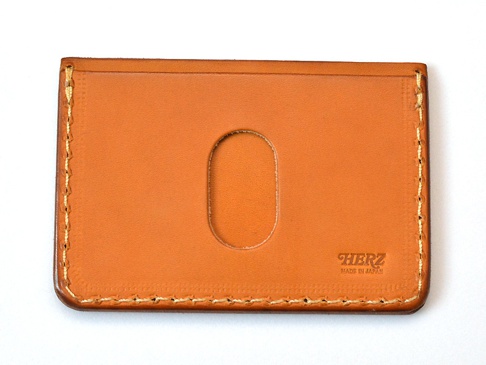 職人手作皮革橫式票券收納夾 (KC-36)，採用光滑的棕褐色皮革製成，正面中央有一個垂直的橢圓形開口。側邊和底部有明顯的米色縫線，右下方壓印有「HERZ MADE IN JAPAN」字樣。