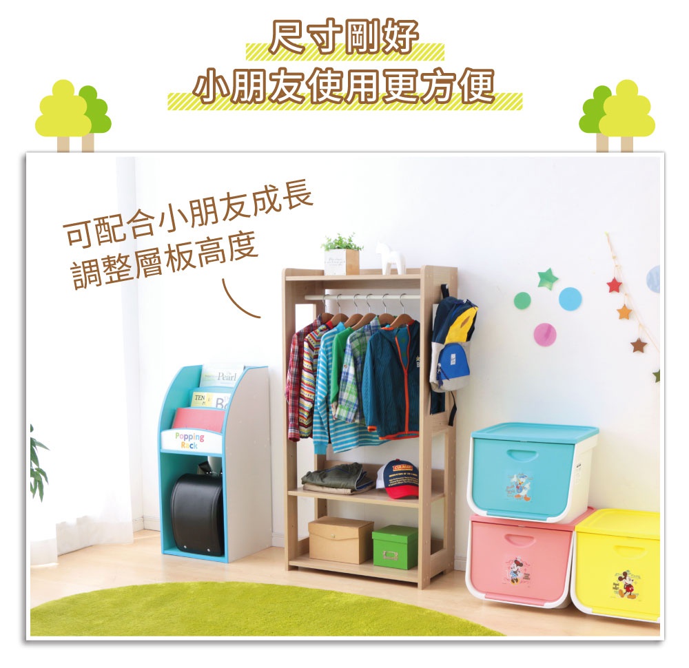 童心木製掛衣架，淺木色，有掛衣桿、兩層置物板，上方掛有彩色衣物，下方置有衣物、帽子和紙盒。旁邊有一個藍白色書架，內有書籍和黑色書包。右側有粉色、藍色、黃色三個卡通圖案的儲物箱。