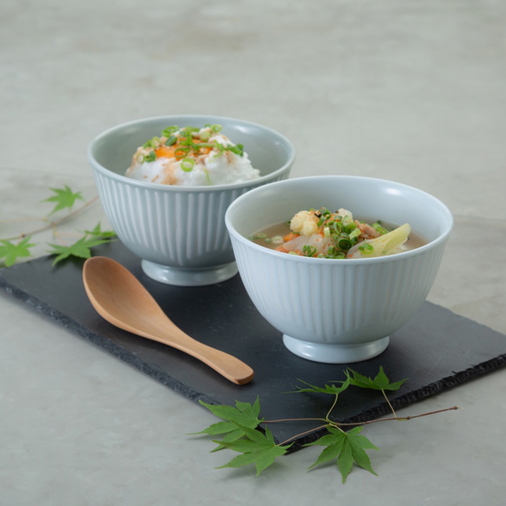 兩個淺藍灰色、帶有垂直紋路的陶瓷飯碗，其中一碗盛有飯和配料，另一碗是湯品。一旁擺放著木製湯匙和綠葉裝飾。