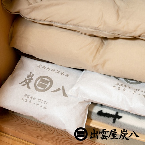 兩個白色包裝袋，上面印有日文「炭八」，標示「室內用調濕木炭」與「高溫炭化 NET 8L 出雲炭素(株)」。包裝袋置於木製層架上，上方疊有米色棉被。